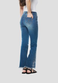 Kék farmer szárbővítéssel, kopott szegéllyel és oldalt virágos hímzéssel. Klasszikus, ötzsebes kialakítással és szabványos derékpánttal rendelkezik.