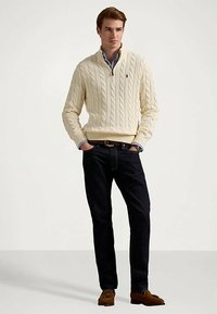 Man wearing cream cable-knit quarter-zip sweater, light blue shirt, dark jeans, brown loafers, standing with hands in pockets on plain background.