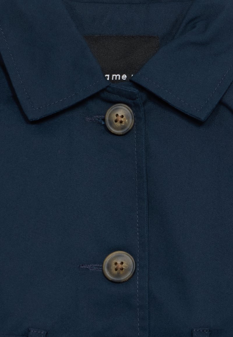 Veste col bleu marine avec une texture lisse. Comprend deux boutons ronds de couleur beige clair et une étiquette sous le col.