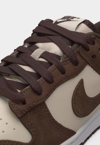 Καφέ και μπεζ αθλητικό παπούτσι Nike με καφέ κορδόνια δεμένα, επενδύσεις σουέτ και ορατό λογότυπο Nike στο πλάι και στη γλώσσα.