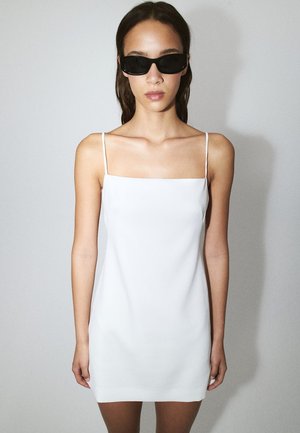 Donna che indossa un mini abito bianco senza maniche e occhiali da sole rettangolari neri, in piedi su uno sfondo grigio chiaro uniforme.