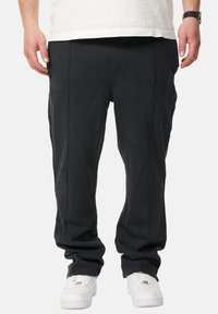 Schwarze, lässig geschnittene Hosen mit sichtbaren Nähdetails und glatter Textur, kombiniert mit weißen Sneakers.