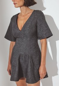 Kratke rokave, temno sivo obleko iz teksturirane tkanine z globokim V-izrezom in zgornjim delom v obliki zvona, ki ima naborke na pasu.