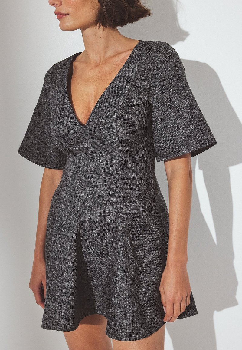 Kratke rokave, temno sivo obleko iz teksturirane tkanine z globokim V-izrezom in zgornjim delom v obliki zvona, ki ima naborke na pasu.