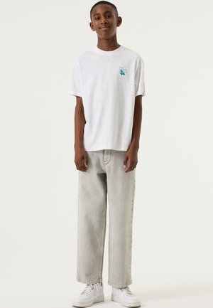 Wit katoenen T-shirt met een kleine blauwe afbeelding op de borst, gecombineerd met losse lichtgrijze spijkerbroek en witte sneakers.