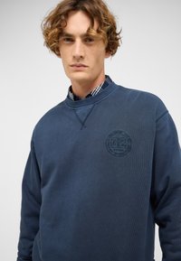 Sweat-shirt bleu marine en tissu doux, avec un patch rond de logo sur la poitrine et un col ras du cou côtelé avec des détails de couture subtils.