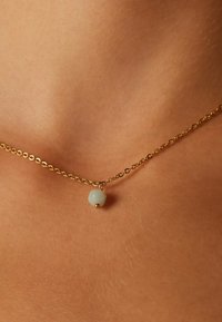 Gouden ketting met een enkele lichtgroene hexagonale kraal, verankerd met een kleine gouden bolaccent. Close-up op de huid.