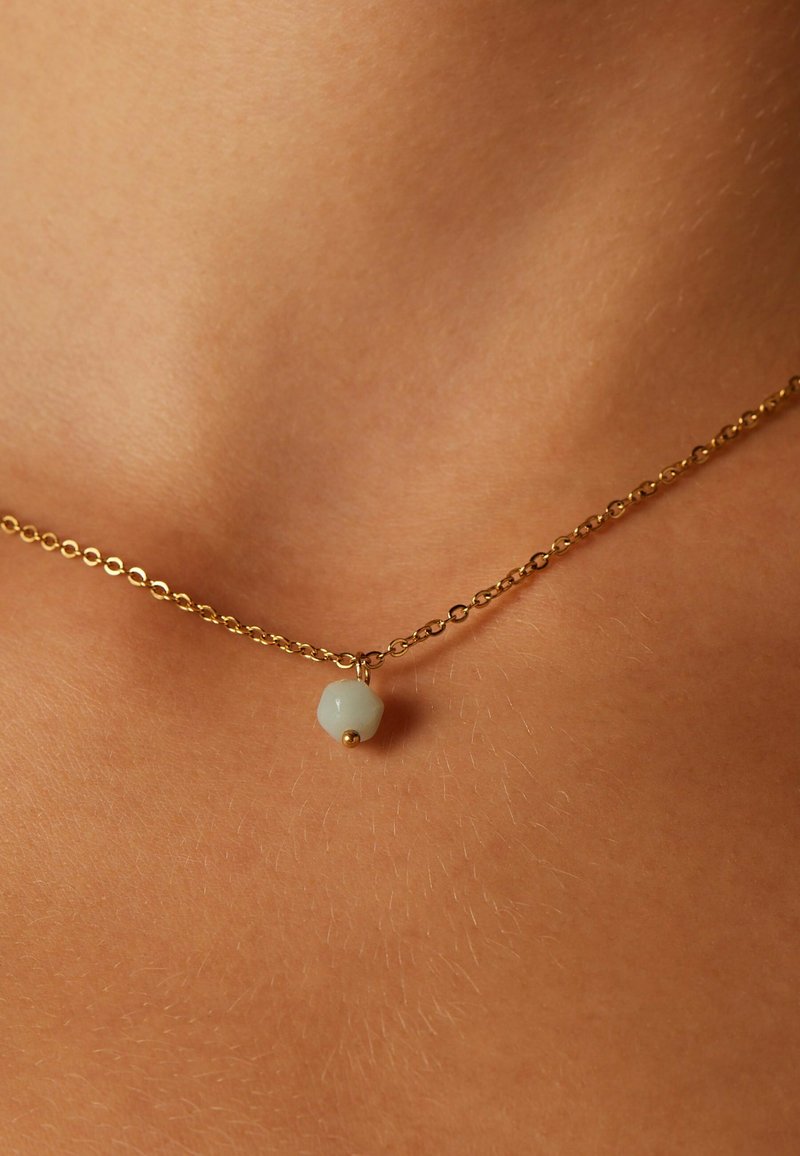 Gouden ketting met een enkele lichtgroene hexagonale kraal, verankerd met een kleine gouden bolaccent. Close-up op de huid.