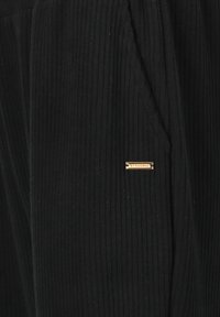 Schwarze Cordhosen mit vertikaler Ripptextur, ausgestattet mit einem kleinen goldenen Markenschild auf der Vorderseite und seitlichen Taschen.