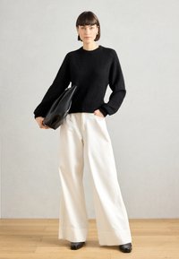 Black wool sweater with a round neckline and loose sleeves, paired with wide white cotton trousers and a black leather bag. Black shoes.