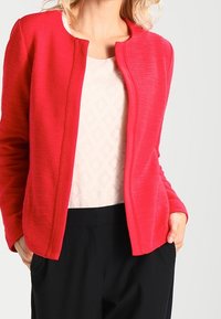 Bright red, textured jacket with a round neckline and no fasteners, worn over a light patterned top and black trousers.