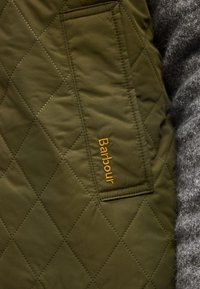 Πανί ελαίας πράσινου με ραφές σε σχήμα ρόμβου και ορθογώνια τσέπη με κεντημένο το "Barbour" με κίτρινο νήμα.