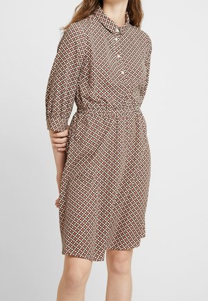 Žena v dlouhých šatech ke kolenům s knoflíky a 3/4 rukávy, s geometrickým vzorem v hnědé, červené a béžové barvě, stojící na jednobarevném pozadí.