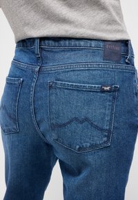 Kék denim farmernadrág klasszikus szabással, hátsó zsebekkel és bőr márkafoglalattal. Az anyag enyhe textúrával és elhalványulás részletekkel rendelkezik.