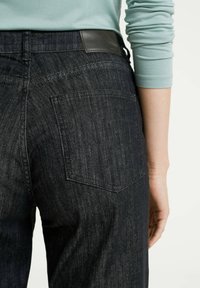 Zadní pohled na osobu oblečenou v tmavě modrých džínech a světle zelené košili s dlouhým rukávem, jejíž rukávy jsou mírně vyhrnuté.