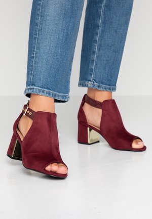 Fødder iført bourgogne ruskind åbne sandaler med blokhæl og ankelremme, kombineret med blå denimjeans, der er cropped over anklerne.