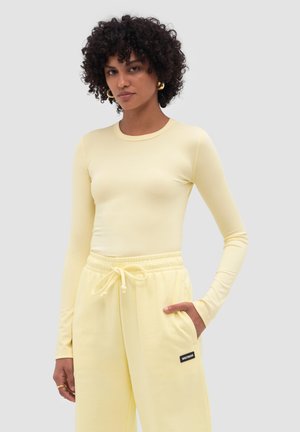 Mujer con cabello rizado que lleva pendientes de aro dorados, una camiseta ajustada de manga larga de color amarillo pálido y pantalones a juego con cordón y detalles en los bolsillos.