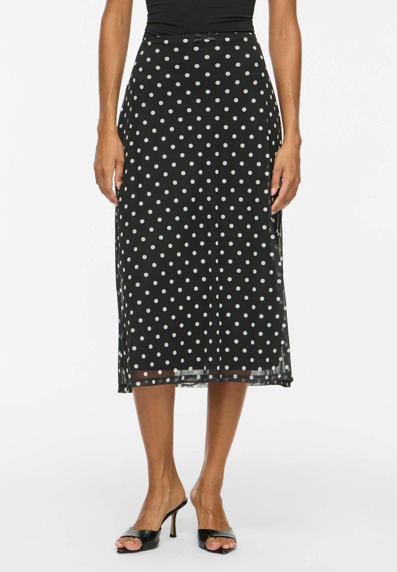 Jupe midi noire à pois blancs, portée avec des sandales à talons ouvertes noires et un haut noir.