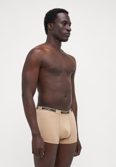 Beige Moschino boxer alsónemű fekete derekú gumival; puha, testhez simuló kialakítás, amely sima anyagot és minimális varrásokat mutat be. A modell oldalról pózol.