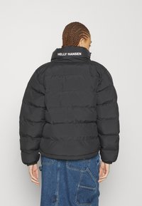 Black puffer jacket with horizontal quilted design, high collar, and white "HELLE HANSEN" logo on the back, made of smooth, insulated material.