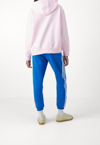 Felpa con cappuccio rosa abbinata a pantaloni da jogging blu con strisce bianche laterali. Il modello indossa sneakers verde chiaro con suola in gomma.