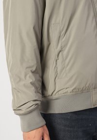 Giacca bomber taupe in tessuto leggero e liscio. Presenta una tasca laterale, polsini a costine e un girovita sagomato.