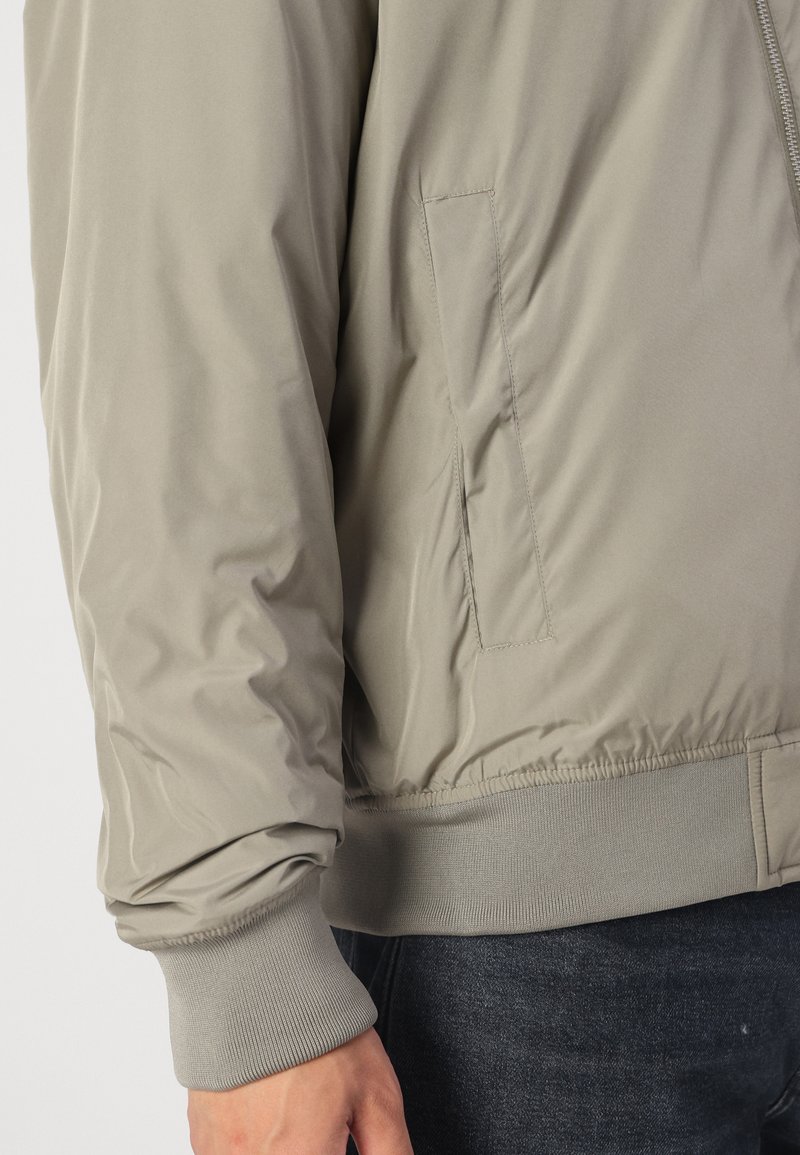 Taupe bomberjack met een gladde, lichte stof. Heeft een zijzak, ribgebreide manchetten en een aansluitende tailleband.