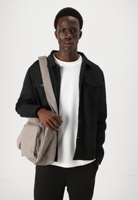 Saco de mensageiro cinza em tela com bolso com fecho, usado como bolsa cruzada. Modelo com uma jaqueta preta de botão sobre uma camisa branca, com calças pretas.