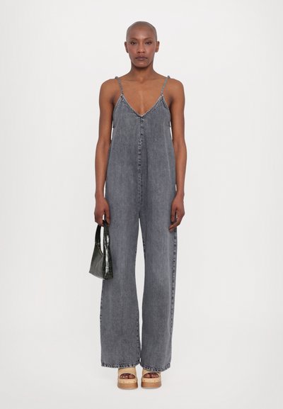 Combinaison en denim grise délavée, design à jambe large, décolleté en V et fines bretelles. Associée à un petit sac à main texturé vert et des chaussures à plateforme.