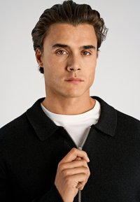 Czarna kurtka rozpinana na suwak z kołnierzem, wykonana z teksturowanej tkaniny, z srebrnym suwakiem, noszona na białej koszulce. Ręka modela trzyma suwak.