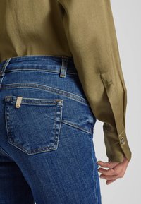 Jeans blu scuro con una texture liscia, caratterizzati da tasche posteriori e cuciture a contrasto, abbinati a una camicia lunga in seta verde oliva.