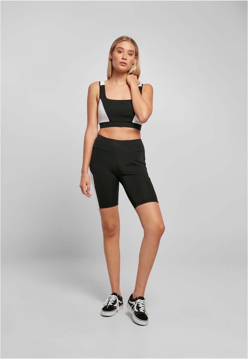 Svart treningscrop-top med hvite sideaksenter og svarte høytalende sykkelshorts; modellen har på seg svarte sneakers, håret ned.