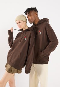 Bruine hoodie met een voorzak en logodetail. Heeft een relaxte pasvorm; gedragen met een beige beanie en een geruite plooienrok.