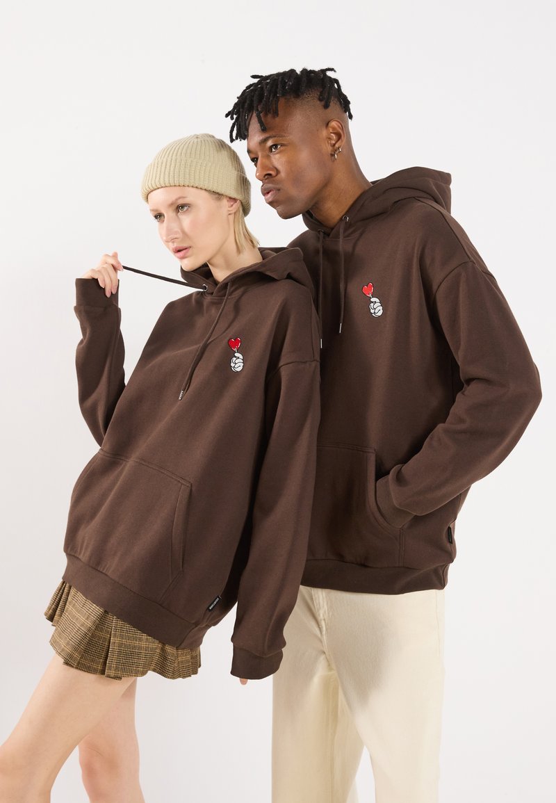 Bruine hoodie met een voorzak en logodetail. Heeft een relaxte pasvorm; gedragen met een beige beanie en een geruite plooienrok.