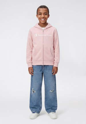 Ragazzo sorridente che indossa una felpa con cappuccio rosa Champion, jeans larghi strappati blu e sneaker bianche, in piedi su uno sfondo chiaro semplice.