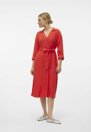 Abito rosso a camicia con cintura annodata, maniche lunghe arrotolate fino ai gomiti e spacco laterale. Indossato con ballerine argento.