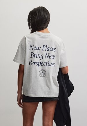 Sötét hajú nő, fehér pólót visel, melyen a "New Places Bring New Perspectives" felirat szerepel, fekete dzsekit tart, háttal a kamerának.