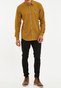 Beżowa koszula z guzikami i małym logo na lewej piersi, w połączeniu z czarnymi, dopasowanymi spodniami i brązowymi butami na sznurówki.