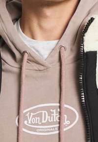 Felpa con cappuccio in taupe chiaro con lacci beige e logo bianco. Indossata sotto una giacca nera con zip e fodera in pile.