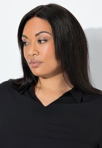 Zwarte blouse met een V-hals. Gladde textuur en aansluitend ontwerp. Model heeft lang steil haar en neutrale make-up.