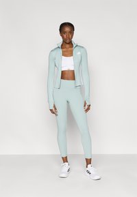 Σετ αθλητικών ρούχων σε ανοιχτό πράσινο μέντας, που αποτελείται από μπουφάν με φερμουάρ και ψηλόμεσα κολάν. Περιλαμβάνει λευκό αθλητικό σουτιέν και λευκά παπούτσια με μαύρες λεπτομέρειες.