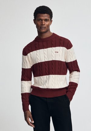Suéter de punto a rayas rojas y crema con patrones de trenzado, cuello redondo y puños acanalados. Pequeño logotipo en el pecho. Usado con pantalones negros.