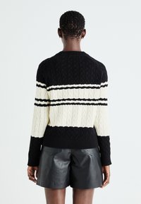 Maglione lavorato a maglia nero e crema con motivo a treccia e righe, dotato di orlo e polsini a coste. Indossato con pantaloni corti in pelle nera.