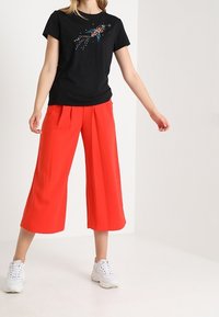 Czarna koszulka z kolorowymi graficznymi detalami, zestawiona z luźnymi czerwonymi spodniami culottes. Obuwie: białe sneakersy. Tło jest jednolite i jasne.