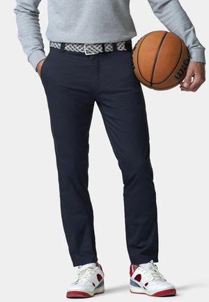 Persoon met marineblauwe broek, riem met patroon, grijs lang mouwen shirt, witte en rode sneakers, houdt met één hand een basketbal vast.