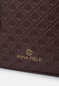 Tumedat pruunid tekstureeritud kunstnahast käekotid, millel on reljeefne lillemuster ja kuldne "ANNA FIELD" logo paremas alaküljel.