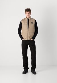 Μπεζ γιλέκο γεμιστό με οριζόντιο ραμμένο σχέδιο, κλείνει με φερμουάρ και έχει όρθιο γιακά, φορεμένο πάνω από ένα μαύρο μακρύ μανίκι πουκάμισο και συνδυασμένο με μαύρο παντελόνι.