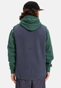 Fleeceväst i mörkgrå, med en grön huva under. Mjuk textur, enkelt mönster och en dragkedja på ryggen.