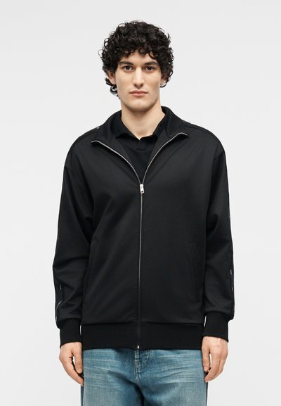 Νεαρός με σγουρά μαλλιά, φορώντας μαύρο μπουφάν με φερμουάρ πάνω από μαύρο πουκάμισο και μπλε τζιν, στέκεται μπροστά σε ένα απλό φωτεινό φόντο.