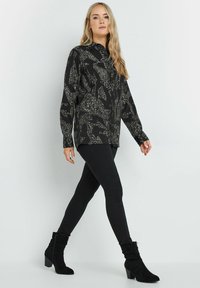 Schwarze gemusterte Bluse mit einem Kragen, langen Ärmeln; kombiniert mit figurbetonten schwarzen Hosen und schwarzen Stiefeletten mit Absatz.